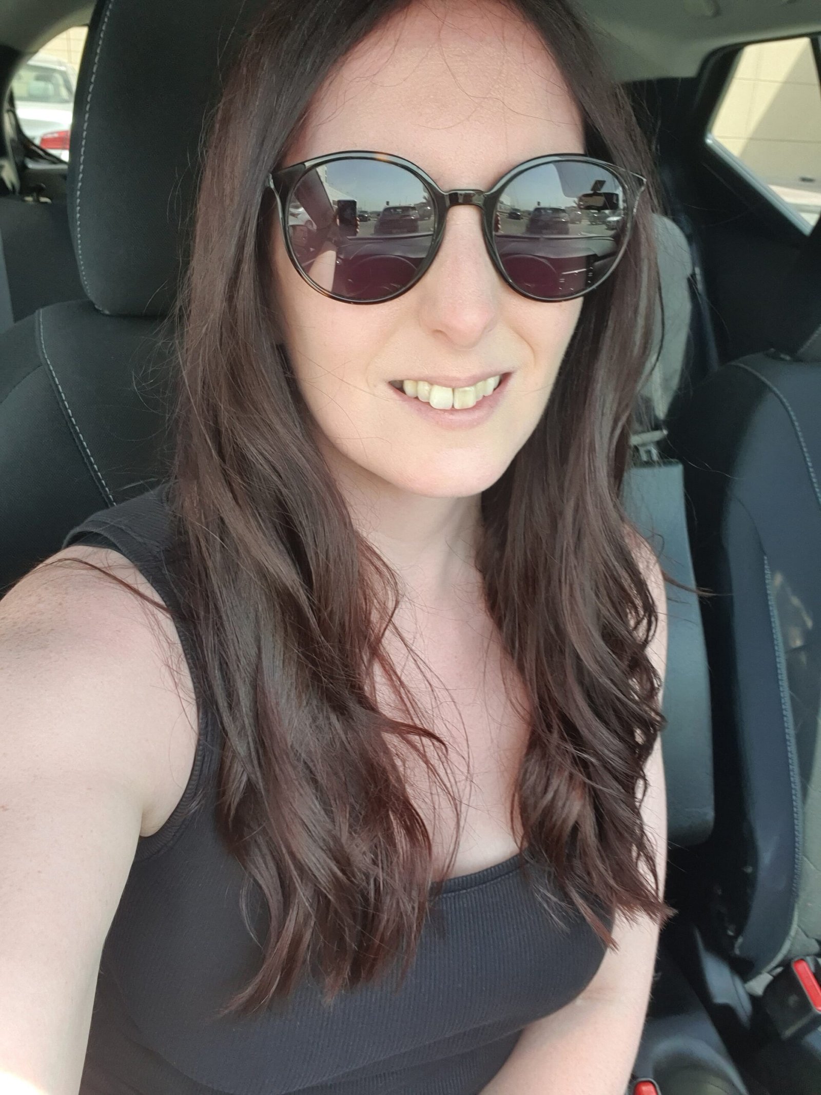 A smiling woman with long dark brown hair takes a close up selfie inside a car. She wears oversized round sunglasses and a black sleeveless top.