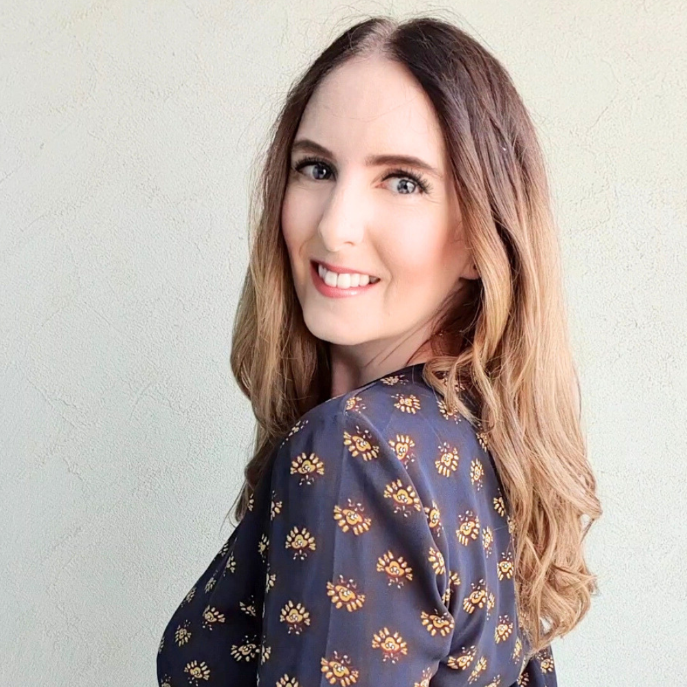 A smiling woman with long brown and blonde hair looks back over her shoulder. She wears a dark blouse with a small gold and orange floral pattern against a pale wall.