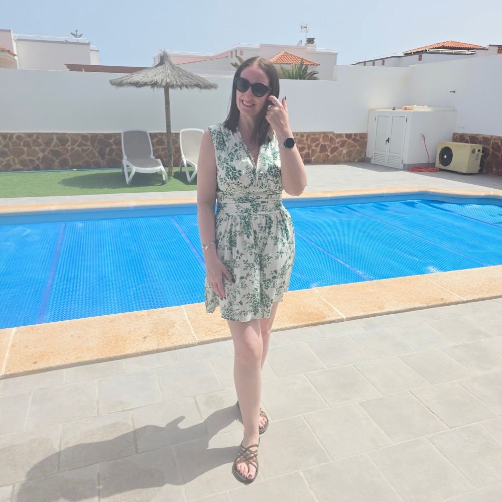 A smiling woman wearing sunglasses and a green floral dress stands beside a bright blue swimming pool. White villas and sun loungers appear behind her under a clear sky.