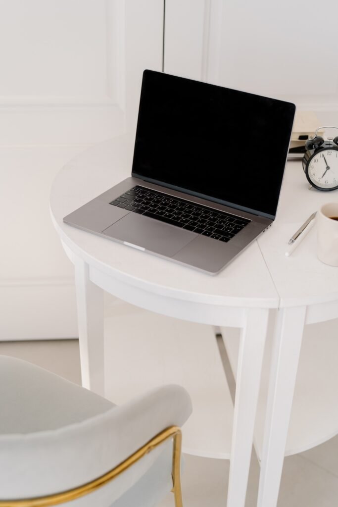 An open silver laptop with a blank black screen sits on a round white desk. A black alarm clock. A white mug. A pen. And part of a pale chair with gold trim complete the minimalist workspace.