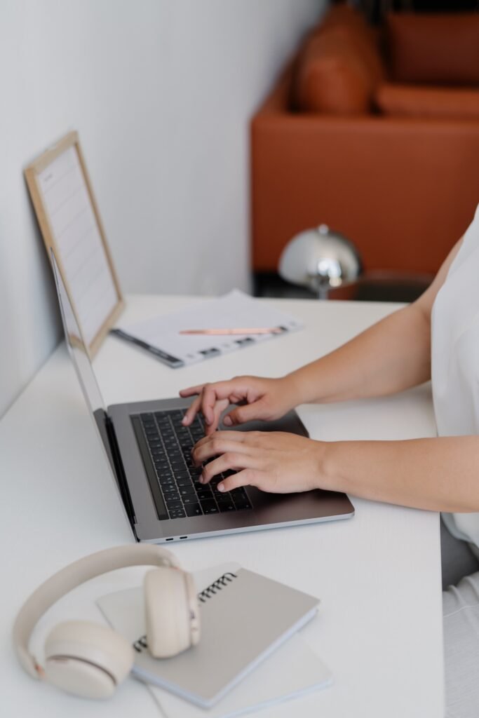 A person types on a laptop at a clean white desk with beige headphones and notebooks in the foreground. A planner and pencil sit beside the computer while a small service bell and warm brown decor appear in the background.