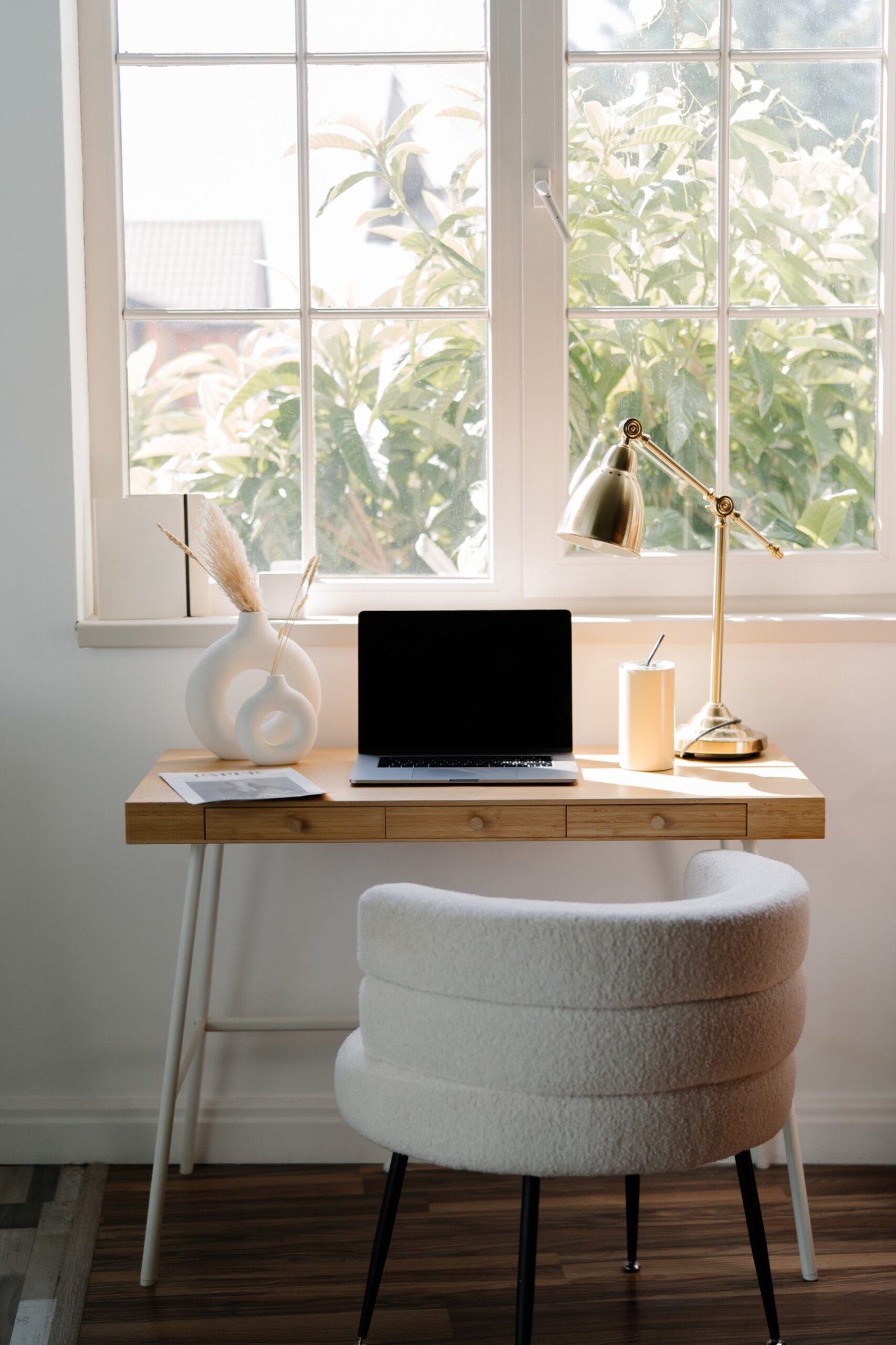 Bright home office with laptop, desk lamp and white chair by a large window